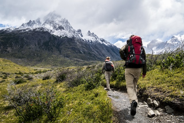 Two walkers on a mountain trail below snowy peaks